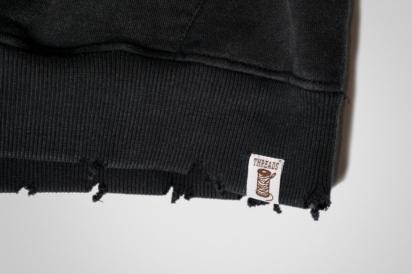 The hem carries a subtle grunge finish, raw yet refined, with a small threads logo stitched in blending edge with identity at the base.